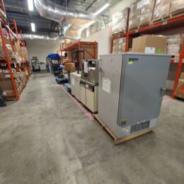 A warehouse with red metal shelves, various boxes, and large industrial equipment lined up on wooden pallets along an aisle with a concrete floor. Ductwork is visible on the ceiling above.